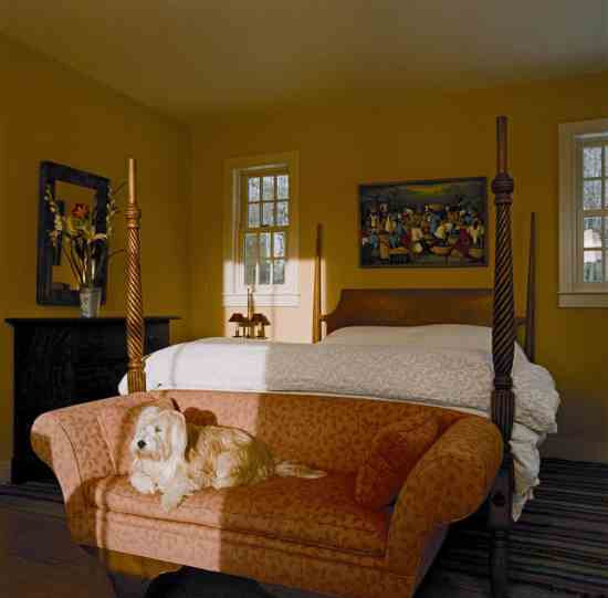 Marguerite basks in the late-afternoon sun on an 18th-century rolled-arm loveseat, another family antique. The bedroom is shielded from electromagnetic fields, and a kill switch turns off all electricity in the room when the couple sleeps. n addition, the all-natural mattress is free of fire-retardant chemicals; linens are organic fibers. The antique, four-poster bed is fitted with a new headboard that expands it to queen size. Above, a colorful painting from Haiti depicts a local fruit market.